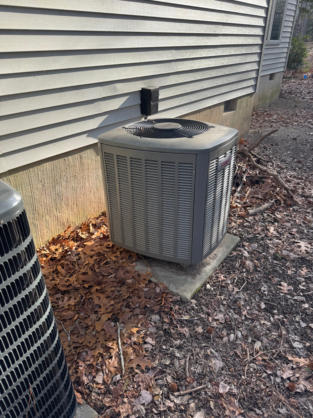Heat pump outdoor unit installed beside a house, surrounded by fallen leaves and natural debris, illustrating compact installation for efficient heating and cooling solutions.
