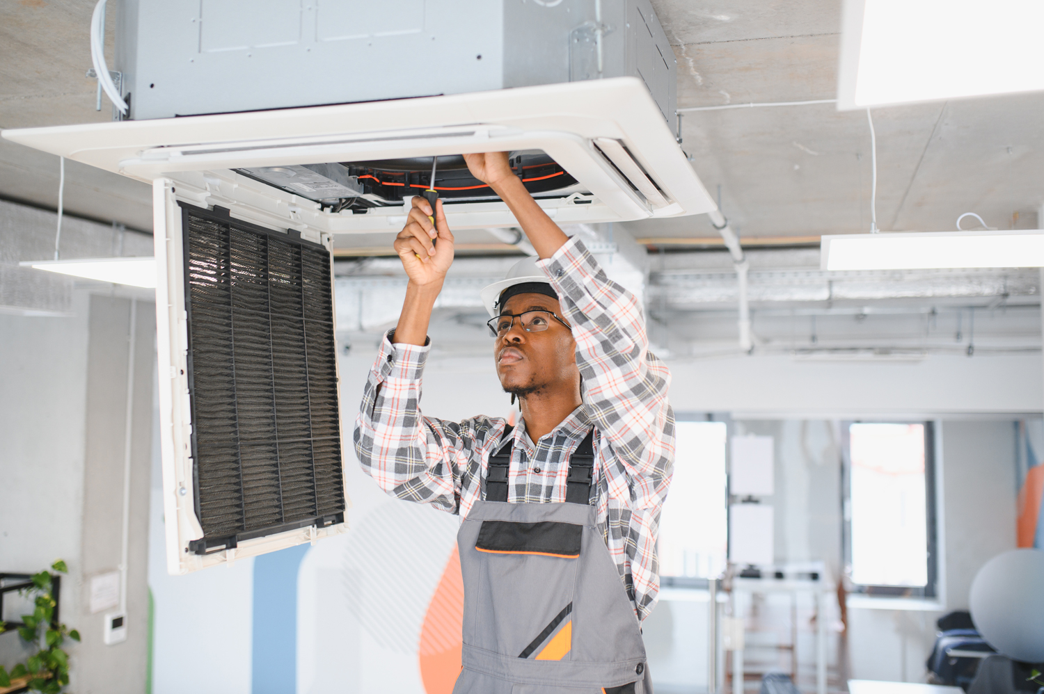 African American technician performing HVAC maintenance on a ceiling-mounted air conditioning unit in a modern office setting, highlighting emergency repair services by PROTECH HVAC LLC.